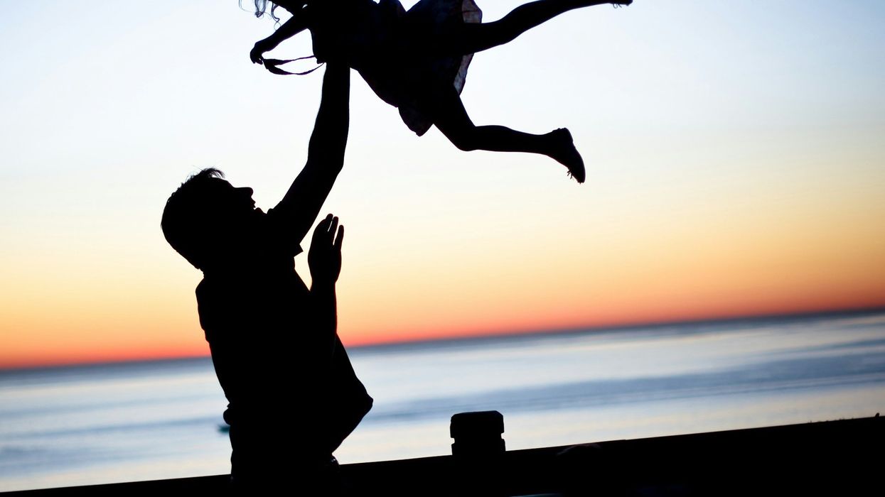 Silhouette photo of a dad joyfully throwing his young daughter in the air while at the beach, at dusk.