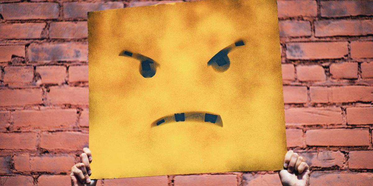 Someone 's hands hold up a tattered yellow sign with a drawn on angry face, against a red brick wall background.