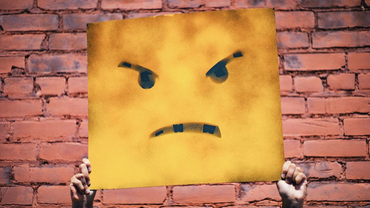 Someone 's hands hold up a tattered yellow sign with a drawn on angry face, against a red brick wall background.