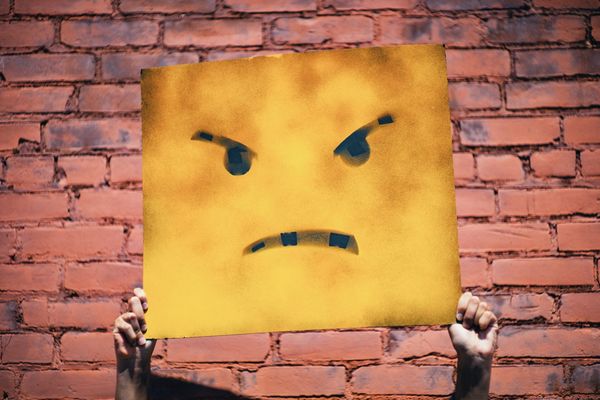 Someone 's hands hold up a tattered yellow sign with a drawn on angry face, against a red brick wall background.