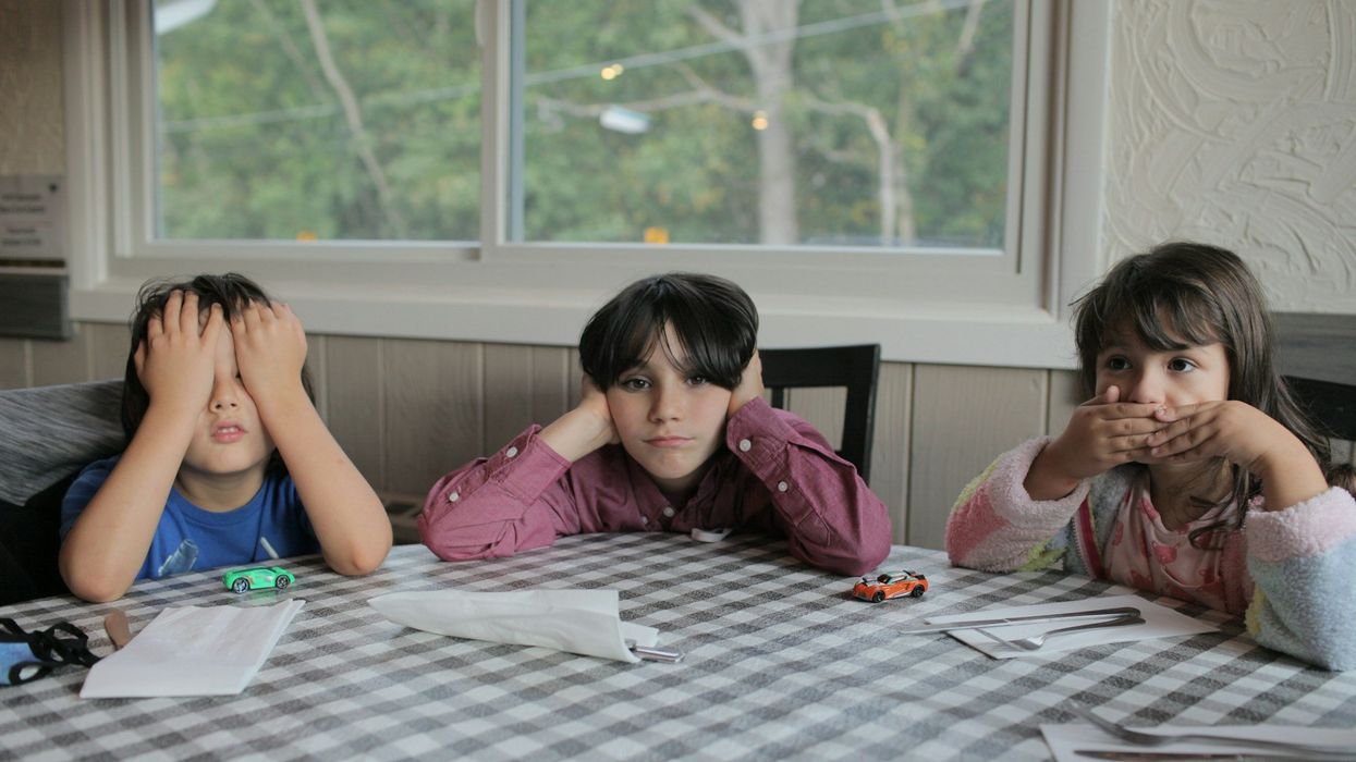 Three children sit at a black and white checkered cloth table. One child covers their eyes, another their ears, the third their mouth.
