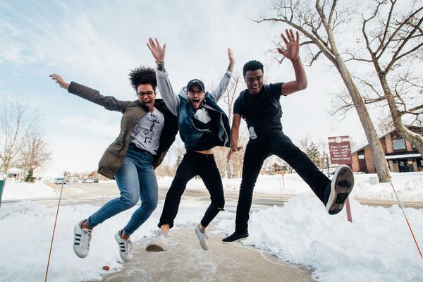 three people jumping on snowy path near bare trees