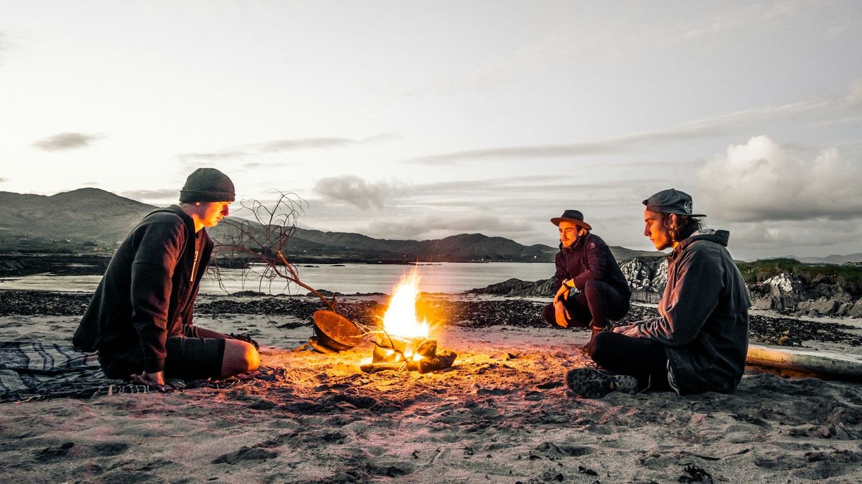 Three young men sit around a campfire on a desolate beach.