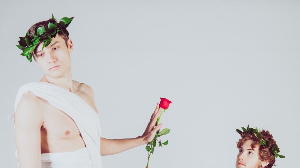 Two attractive, young men pose in togas against a white backdrop. One is on bended knee giving the either a rose. The man standing rejects the rose.