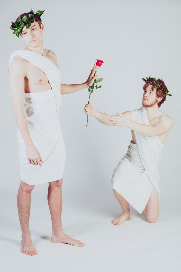 Two attractive, young men pose in togas against a white backdrop. One is on bended knee giving the either a rose. The man standing rejects the rose.
