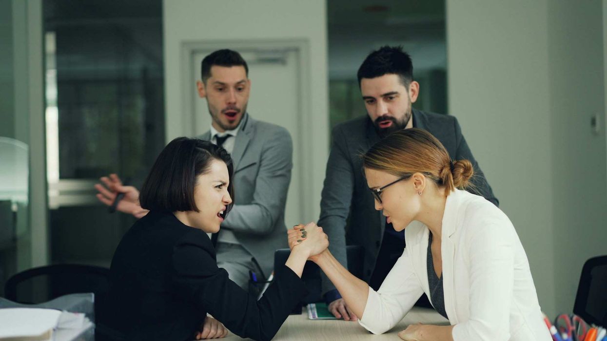 Two female co-workers arm wrestle while two male co-workers look on in shock.