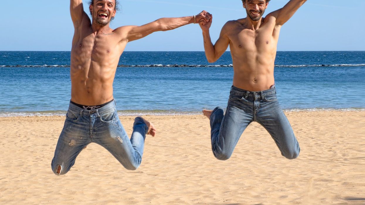 Two young, muscular men jump in the air holding hands, on a beach. Both are shirtless and are wearing light blue jeans