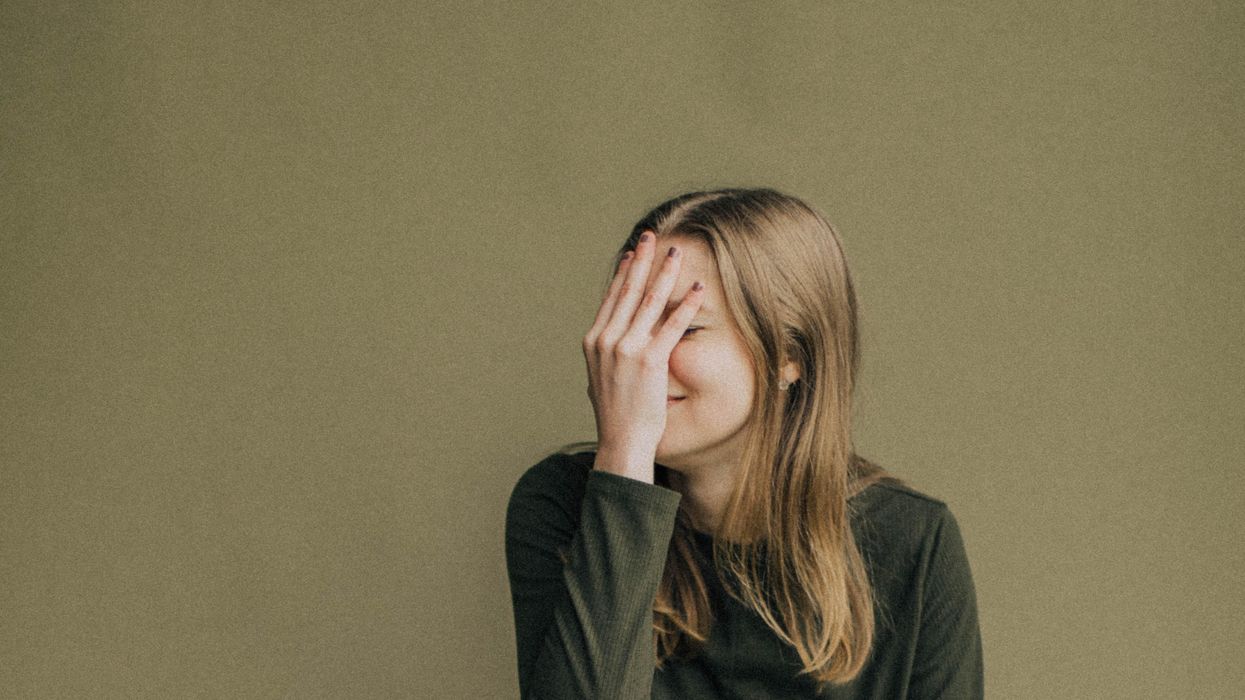 woman with long blonde hair in a green long sleeve shirt with her hand covering her face